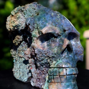 以下が含まれることがあります： テクスチャ加工された表面を持つ、彫刻された石の頭蓋骨の彫刻。 頭蓋骨は主に青と緑で、紫と茶色のヒントがあります。 歯は顎のラインに彫られています。 彫刻はぼやけた緑の背景に対して表示されます。