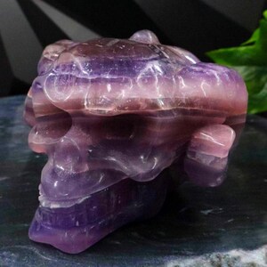 Può includere: Una scultura a forma di teschio scolpita in cristallo di fluorite viola e rosa a strati. Il teschio presenta dettagli e una superficie lucida, che mostra le variazioni naturali di colore della pietra. Un oggetto decorativo unico.