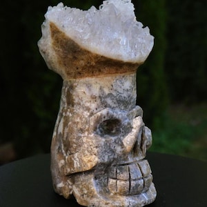 Pode incluir: Uma escultura de crânio em pedra esculpida com uma coroa de cristais de quartzo transparentes. O crânio é detalhado com órbitas oculares, um nariz e uma boca sorridente. A pedra tem uma mistura de cores cinza, marrom e branco.