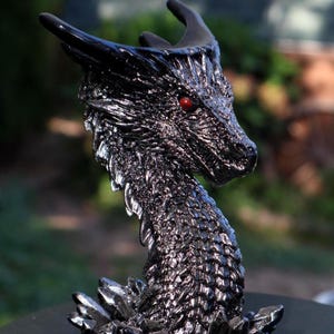 May include: A detailed black dragon figurine with red eyes. The dragon has textured scales and horns, and is perched on a base of jagged crystals. The sculpture is highly detailed and appears to be made of a metallic material.