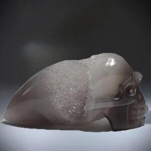May include: A carved crystal skull in a pale purple hue, with a smooth, translucent finish. The top of the skull has a rough, crystalline texture. The teeth and eye sockets are well-defined, showcasing intricate detailing.