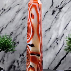 May include: A tall, polished agate crystal point with a striking pattern of swirling orange, white, and black bands. The crystal is displayed on a dark surface against a marble backdrop, showcasing its natural beauty.