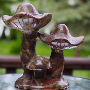 May include: A carved stone sculpture of three mushrooms in shades of brown, red, and green. The mushroom caps have a curved shape with detailed gills. The sculpture is a decorative item, likely for home decor.