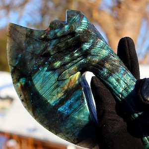Puede incluir: Una cabeza de hacha tallada a mano en piedra labradorita. La hoja tiene un borde curvo y el mango presenta un diseño detallado. La piedra muestra una mezcla de colores azules, verdes y dorados. El hacha es sostenida por una mano enguantada.