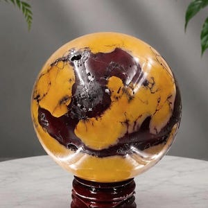 May include: A polished spherical stone featuring a marbled pattern of mustard yellow and deep maroon with dark, thin, vein-like inclusions throughout the surface. The sphere is displayed resting on a circular, dark brown wooden stand with a tiered base.