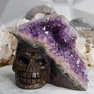 May include: A carved brown skull sculpture with a large amethyst geode on top. The amethyst features deep purple crystals. The skull has a detailed, smiling face. The sculpture is on a white surface.