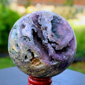 May include: A spherical, polished geode with a complex, textured surface. The geode features a mix of purple, blue, and cream colors, with a cavity revealing crystalline formations. It is mounted on a red wooden stand.