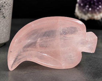 May include: A polished rose quartz crystal carved into the shape of a leaf. The stone features a translucent, pale pink hue with natural internal inclusions and a smooth, glossy surface finish, showcasing a curved, pointed tip and a stem.