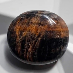 May include: A spherical gemstone, polished to a high sheen, displaying a blend of dark brown and orange tones. The surface features a distinctive pattern of lines and swirls, giving a marbled appearance. This gemstone is a decorative item.