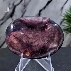 May include: A rounded, polished gemstone in shades of deep purple and burgundy. The stone is displayed on a clear acrylic stand, highlighting its smooth, reflective surface. The background features a marble pattern and a touch of greenery.