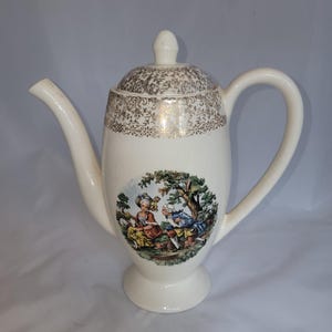 May include: A cream-colored ceramic coffee pot with a lid, featuring a gold filigree pattern around the top rim and a central circular decal depicting two figures in period clothing sitting in a landscape with trees and greenery.