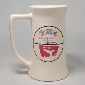 Puede incluir: Jarra de cerveza de cerámica color crema con asa. La jarra presenta un diseño circular con las banderas de Estados Unidos, las Naciones Unidas y Corea del Sur. El diseño incluye el texto "CHINCHON-NI A.O.M", un mapa rojo y el texto "PY-Do" y "DMZ".