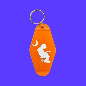 Puede incluir: Llavero de motel de plástico naranja con una silueta blanca de una persona sentada en un taburete con una luna creciente en el fondo.