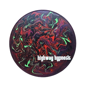 Puede incluir: Un disco de vinilo negro con un diseño de pintura rojo, verde y morado en espiral. El texto "highway hypnosis" está impreso en blanco en el disco.