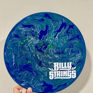 Può includere: Un oggetto decorativo rotondo, blu e verde, con un motivo a spirale. L'oggetto presenta il testo bianco "Billy Strings". L'oggetto è tenuto da una mano.