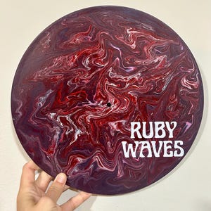 Pode incluir: Um disco de vinil vermelho, branco e roxo com o texto "RUBY WAVES" em letras brancas.