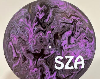 May include: A round, black and purple decorative item with a swirling, marbled design. The word "SZA" is printed in white block letters. The item is held by a hand.