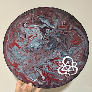 Puede incluir: Un disco de vinilo circular con un diseño de pintura marmolada en tonos rojo, azul claro, negro y blanco. La superficie muestra un patrón fluido y arremolinado, con un símbolo gráfico blanco impreso en el cuadrante inferior derecho.