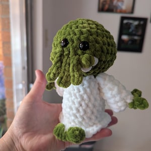May include: A small, handmade crochet plush toy featuring a round, olive green head with dangling tentacles, two black plastic eyes, and a white, textured body with green hands and feet, crafted from soft, thick yarn in a plush, fuzzy texture.