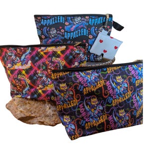 May include: Three zippered pouches featuring colorful graphic prints of cats riding dinosaurs with the text "MONGO IS APPALLED!" and "THIS IS AN OUTRAGE". One pouch includes a small white square charm with red hearts attached to the zipper pull.