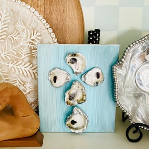 May include: A blue painted wooden board with five oyster shells arranged in a cross shape. The shells are natural and have a white and gray color.