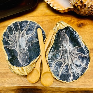 Può includere: Due piatti a forma di conchiglia di ostrica in nero e oro con un motivo di polpo bianco. I piatti sono abbinati a due cucchiai di legno.