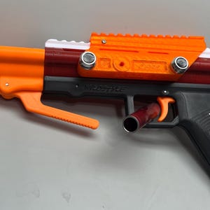 Puede incluir: Una pistola de juguete de color naranja brillante y negro con un cañón rojo y blanco. La pistola tiene un mango negro y un gatillo plateado. La pistola tiene una mira negra y naranja en la parte superior.