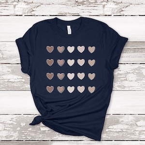 May include: Navy blue t-shirt featuring a pattern of heart shapes in various shades of brown and beige. The hearts are arranged in a grid pattern. The shirt has short sleeves and a crew neck. The fabric appears to be soft.
