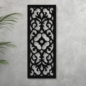 May include: Black rectangular wall panel with an intricate, symmetrical floral design. The panel is made of a solid material and is mounted on a light gray wall. The design features swirling patterns and a central flower-like motif.