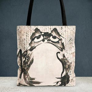 May include: Tote bag featuring a black and white illustration of a frog with a sad expression. The frog is drawn in a traditional Japanese style, with calligraphy script in the background. The bag has black handles.
