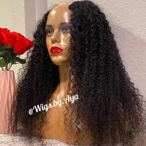 May include: A mannequin head with a dark curly wig. The mannequin has red lipstick and eye makeup. A pink vase with red roses is to the left. The text "@Wigs.by.Aya" is on the image.