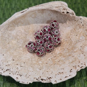 May include: A collection of pink rhinestone beads in a white shell-shaped dish. The beads are silver-toned and have a sparkly finish.