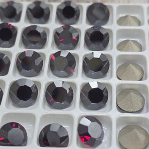 May include: Close-up of a tray filled with dark gray, faceted rhinestones. The rhinestones have a circular shape with multiple facets, reflecting light. Some rhinestones have a hint of red. Also included are a few silver-colored, pyramid-shaped stones.
