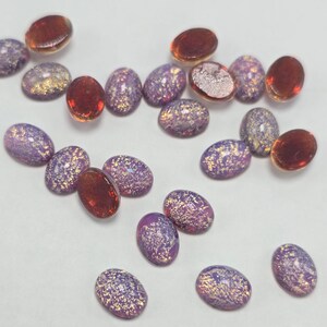 May include: A collection of oval-shaped cabochons featuring a mix of translucent deep amber-colored glass and opaque purple glass with embedded gold-toned metallic flakes. The stones are smooth, rounded, and display a reflective, speckled surface.