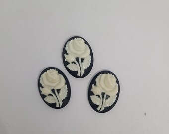 May include: Three white cameo-style rose flower embellishments with a black background. The roses are in an oval shape.