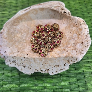 May include: A collection of gold-colored, ornate beads with embedded red and clear gemstones. The beads are arranged inside a natural, off-white seashell, set against a green woven background. The beads have a vintage or antique aesthetic.