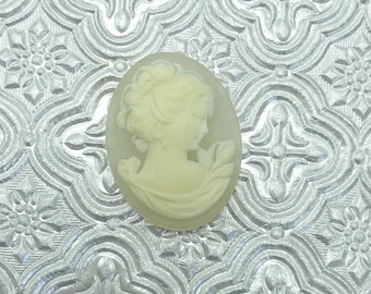 May include: A white cameo featuring a woman's profile with a detailed hairstyle and a delicate floral design. The cameo is oval-shaped and has a smooth, polished finish.