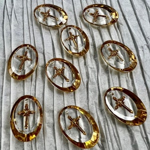 May include: Nine clear oval-shaped pendants with gold-colored cross designs. Each pendant has a gold-colored border and a small hole at the top for stringing. The pendants are arranged on a textured, light gray surface.