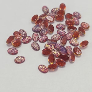 May include: A collection of small, oval-shaped glass cabochons featuring a mix of translucent amber, deep orange, and iridescent purple hues. Each piece displays a textured, metallic-flecked surface that catches the light, creating a shimmering effect across the scattered assortment of smooth, polished decorative stones.