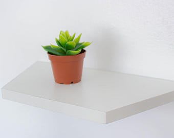 May include: A small, artificial succulent plant in a brown pot sits on a white, angled shelf. The shelf is mounted on a white wall. The plant has bright green leaves, and the pot is a standard terracotta color.