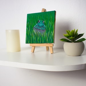 May include: A small painting on a wooden easel depicts a green frog sitting on a rock in a field of green grass. The painting is on a white shelf with a white candle and a potted succulent plant.