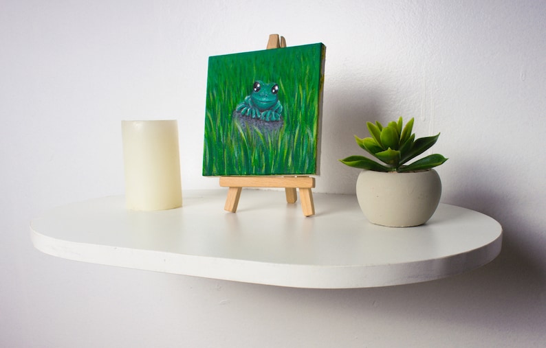 May include: A small painting on a wooden easel depicts a green frog sitting on a rock in a field of green grass. The painting is on a white shelf with a white candle and a potted succulent plant.