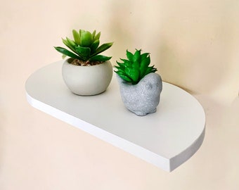 May include: A white, half-round shelf with rounded edges, mounted on a wall. Two small potted plants, one in a gray bird-shaped planter and the other in a round gray planter, are displayed on the shelf.