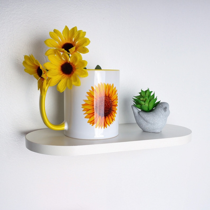 May include: White ceramic mug with a yellow sunflower design on a white oval shelf with a small gray bird figurine and a small green plant.