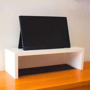 May include: A white monitor stand with a black computer monitor and keyboard. The stand is rectangular with an open space underneath for the keyboard. The monitor is angled back, and the keyboard is black.