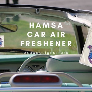 Puede incluir: Un ambientador de coche Hamsa con un diseño azul y rosa. El ambientador está colgado de un espejo retrovisor en un coche. El texto "HAMSA CAR AIR FRESHENER" y "expodesignsstore" son visibles en la imagen.