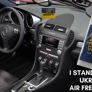 May include: A blue and yellow car air freshener with the text "I Stand With Ukraine" and a QR code. The air freshener is hanging from the rear-view mirror of a car.