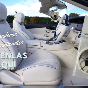 May include: A white car interior with a white leather seat and a black and white football air freshener hanging from the rearview mirror. The air freshener is packaged in a white plastic bag with the text "EXP Designs Air Fresheners". The text "Banderas Aromatizantes Obténlas Aquí" is visible on the seat.