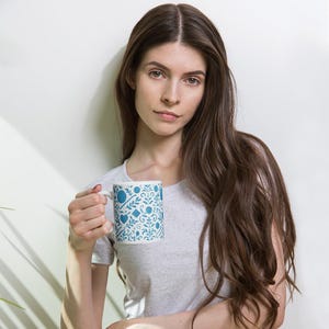 May include: A white ceramic mug with a blue and white floral pattern. The mug features a repeating design of stylized flowers, hearts, and geometric shapes. The mug is held by a person.