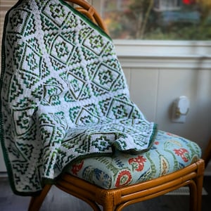 以下が含まれることがあります： 幾何学模様の白と緑のクロシェの毛布が、花柄のクッションが付いた木製の椅子にかけられています。